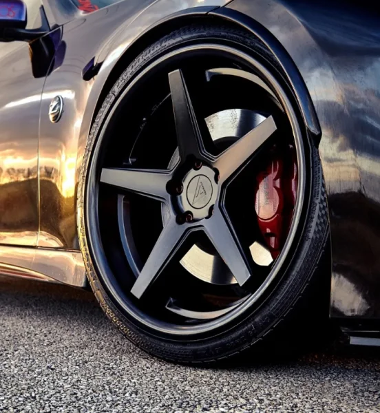 Close-up of a lowered black sports car featuring large matte black 5-spoke wheels with low-profile tires and red brake calipers, reflecting the sunset light on the vehicle’s glossy body.