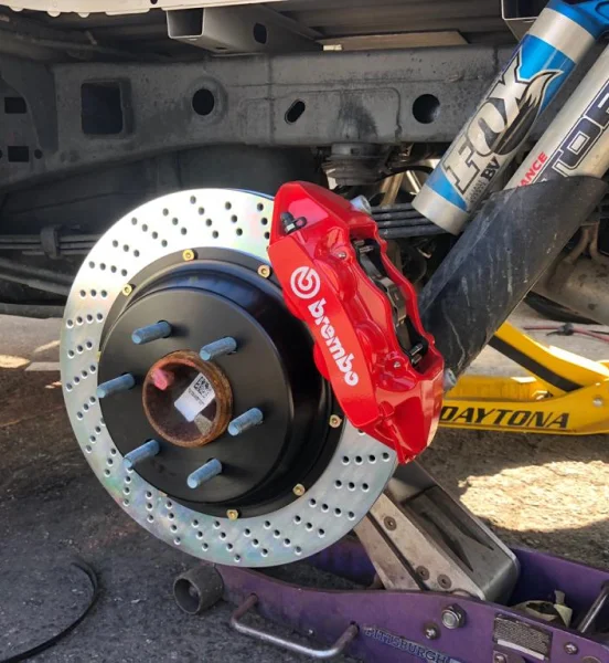 Close-up of a vehicle’s suspension and braking system during installation, showcasing a red Brembo brake caliper mounted on a drilled and slotted rotor, with a FOX shock absorber and jack stands visible in the background.