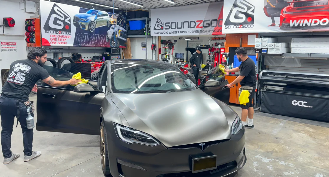Technicians at Soundz Good install window tint on a matte black Tesla Model S inside a detailed auto shop. The workshop features wall banners, tire racks, and various automotive tools, showcasing the business’s focus on high-end customization and professional service.