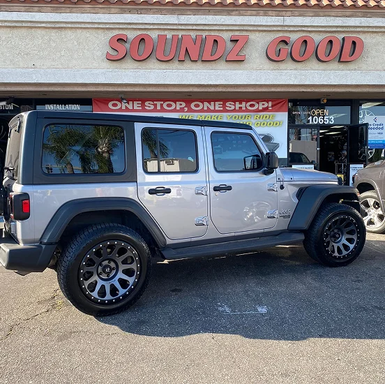 A silver Jeep Wrangler with black accents and custom black wheels parked in front of a store named "Soundz Good," which offers vehicle audio and installation services. The storefront features signs reading "ONE STOP, ONE SHOP!" and "WE MAKE YOUR RIDE SOUND GOOD!"