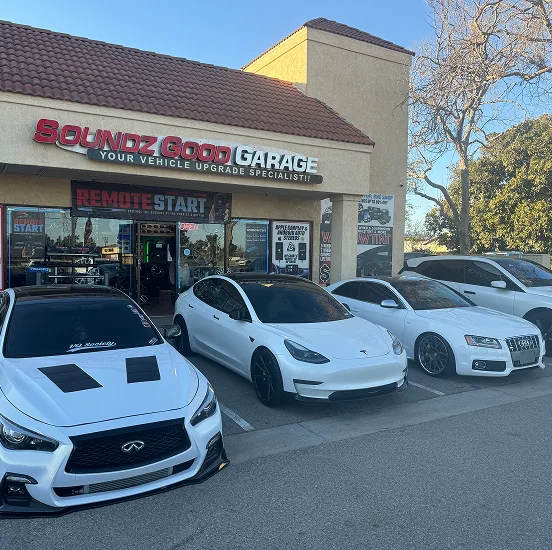 Several customized white vehicles, including an Infiniti, Tesla Model 3, and Audi, are parked outside "Soundz Good Garage," a vehicle upgrade shop with signage advertising remote start, audio systems, and other customizations.