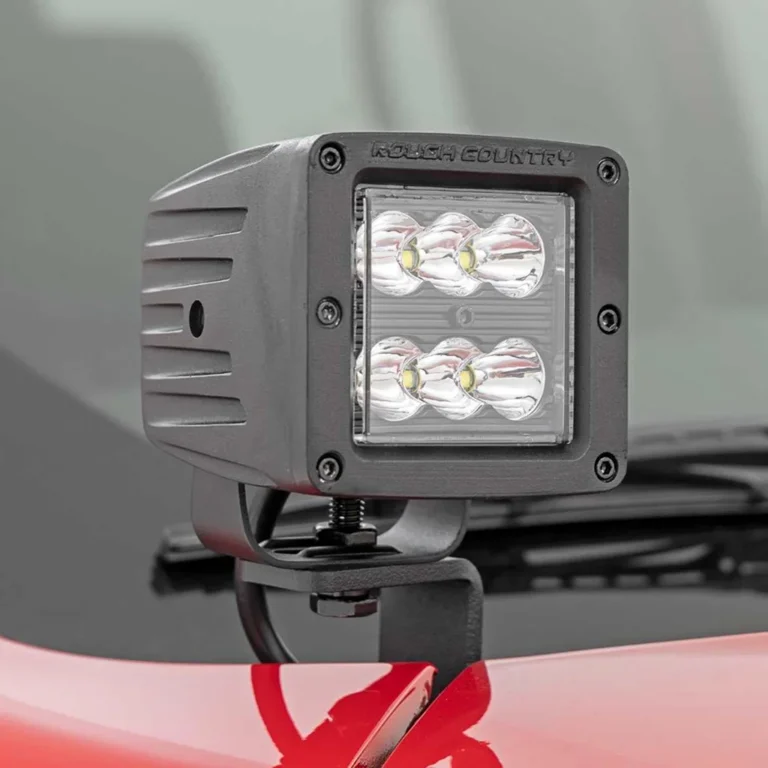 Close-up of a Rough Country LED pod light mounted on the hood of a red vehicle. The light features six LED lenses arranged in two rows of three, housed in a durable black metal casing with visible mounting hardware.