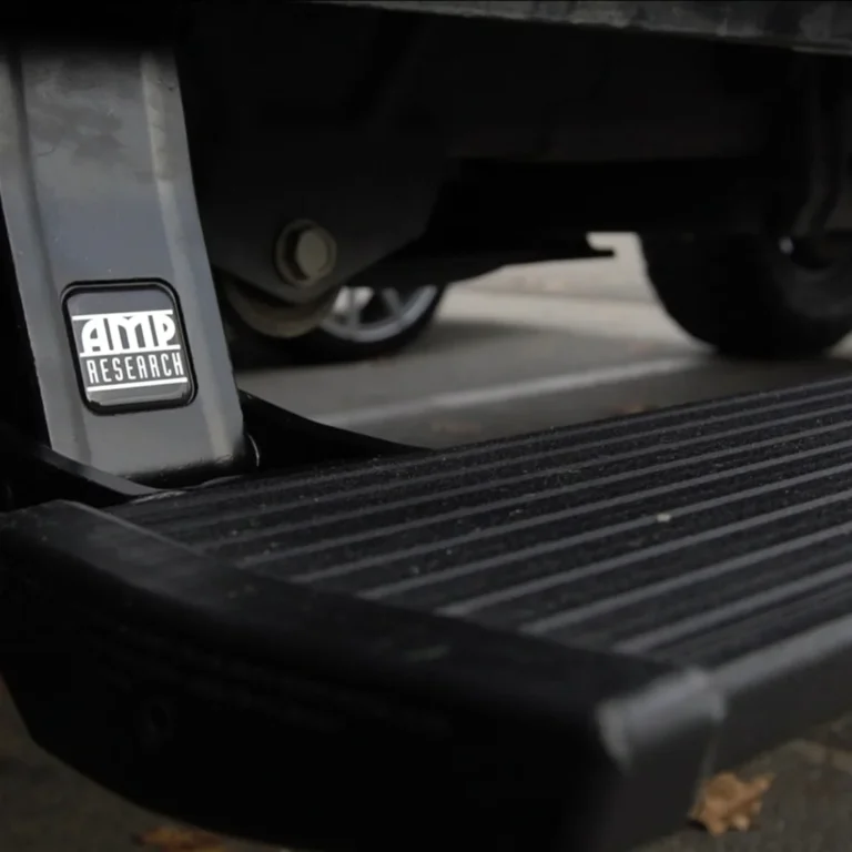 Close-up of an AMP Research retractable running board on a truck, showing the logo on the mounting bracket and the textured step surface designed for secure footing.