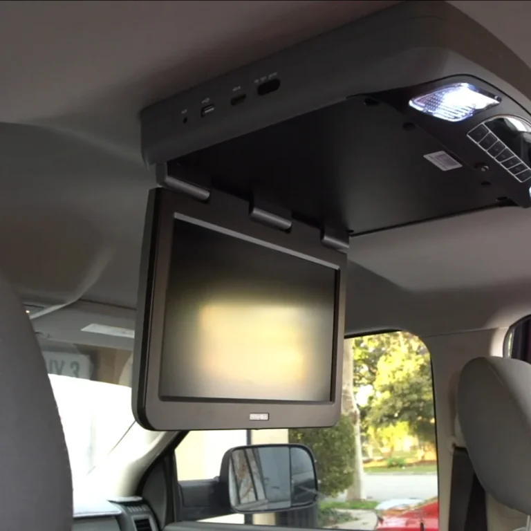 Ceiling-mounted flip-down monitor installed in a vehicle interior, positioned above the backseat area. The screen is off, and overhead controls with input ports and a built-in light are visible on the housing unit.