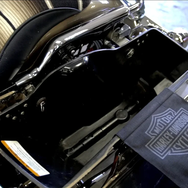 Open Harley-Davidson motorcycle saddlebag showing an empty black interior with visible hardware and a Harley-branded cloth partially covering the lower right corner. A custom seat with a striped cover is also visible above.