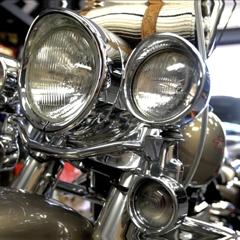 Close-up of a vintage-style motorcycle front end featuring triple chrome headlights and a spotlight, with a rolled blanket strapped above the headlight assembly. The chrome detailing reflects the workshop environment.