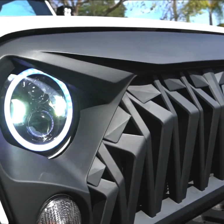 Close-up of a custom Jeep front end featuring a matte black aggressive grille with angular design and a halo-style LED headlight emitting bright white light, paired with a lower auxiliary light.