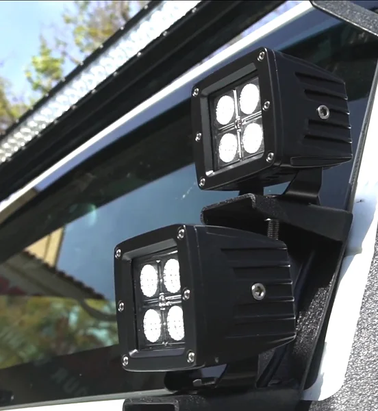 Close-up of two black LED off-road pod lights mounted on the side of a vehicle near the windshield, each housing four bright LED bulbs in a square pattern, with a light bar visible on the roof above.
