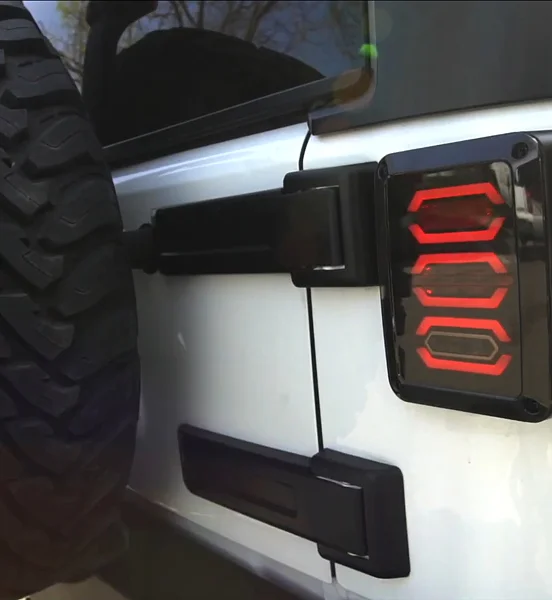 Close-up of a rugged Jeep rear corner showing a custom red LED tail light with a geometric hex pattern, mounted next to a large spare tire and black tailgate hinges on a white vehicle.