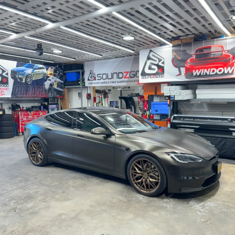 A matte black Tesla Model S with bronze aftermarket wheels parked inside the Soundz Good garage, surrounded by banners, tools, and equipment for custom vehicle upgrades including window tint, audio, and suspension services.
