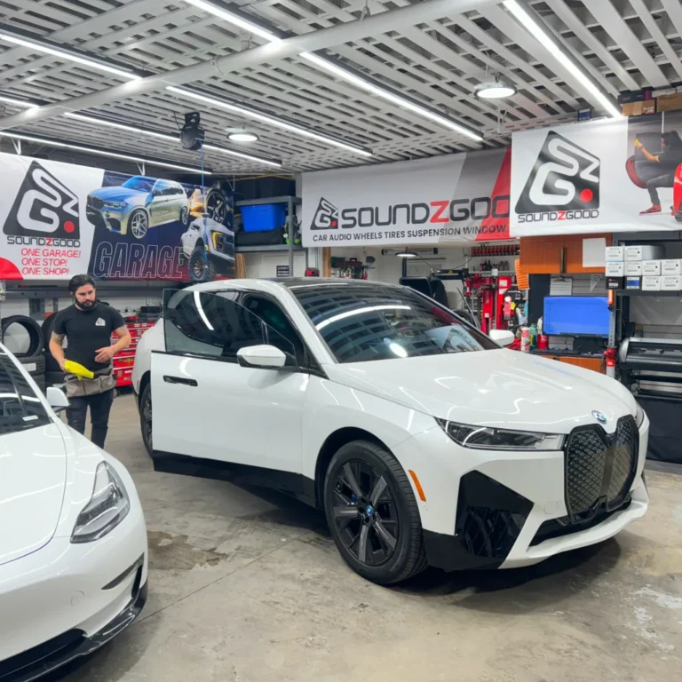 A white BMW iX electric SUV inside the Soundz Good garage, with a technician nearby holding detailing cloths. The vehicle is positioned next to a white Tesla Model 3, surrounded by banners and custom car upgrade tools in a professional workshop setting.