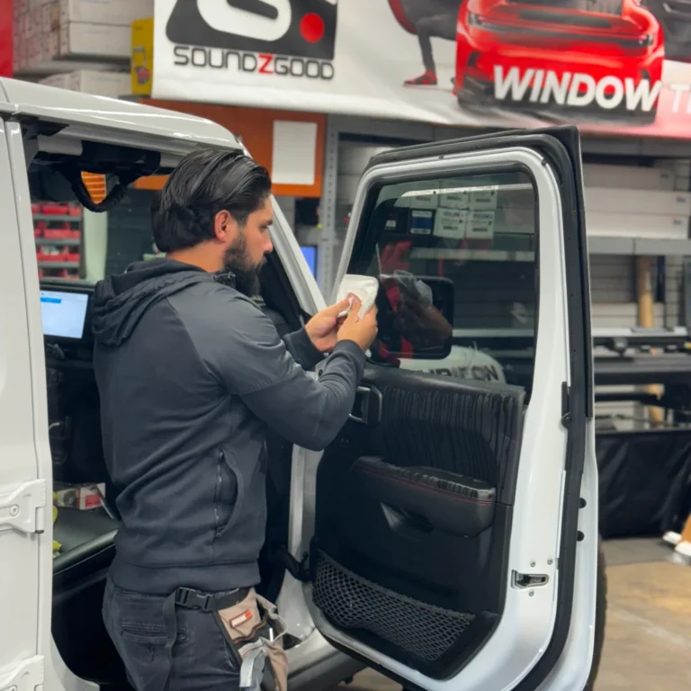 Technician at Soundz Good applying window tint film to the rear passenger door of a white Jeep Wrangler. The workspace features professional tools, banners promoting window tint services, and an organized automotive upgrade area.