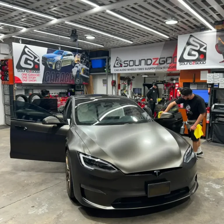 A matte black Tesla Model S being detailed at Soundz Good garage, with technicians working on window tint installation. The workshop is equipped with tools, banners, and custom vehicle upgrade equipment, emphasizing the shop’s focus on performance and aesthetics.