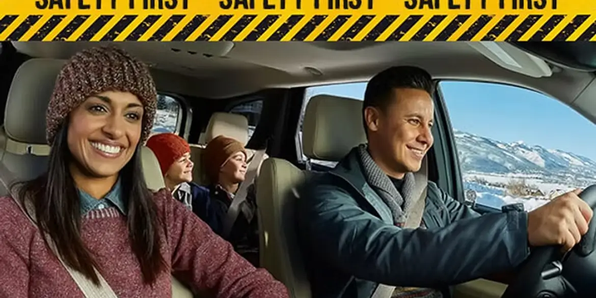 Smiling family of four wearing winter clothing rides safely buckled up in a car on a snowy mountain road, with a “SAFETY FIRST” banner in black and yellow stripes at the top.