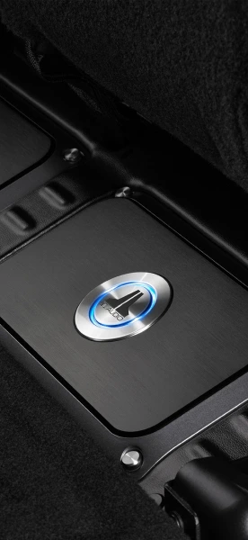 Close-up of a Focal car audio subwoofer with a brushed metal finish and illuminated logo embedded in a sleek black housing, installed in a vehicle’s trunk or under-seat area.