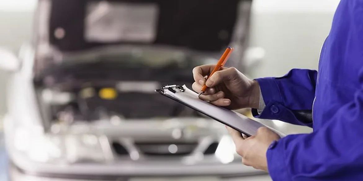 A mechanic wearing a blue uniform writes on a clipboard in front of a car with its hood open, indicating an inspection or maintenance check.