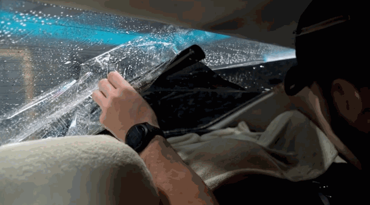 Close-up of a technician peeling off old window tint from the inside of a rear windshield, revealing the glass beneath.