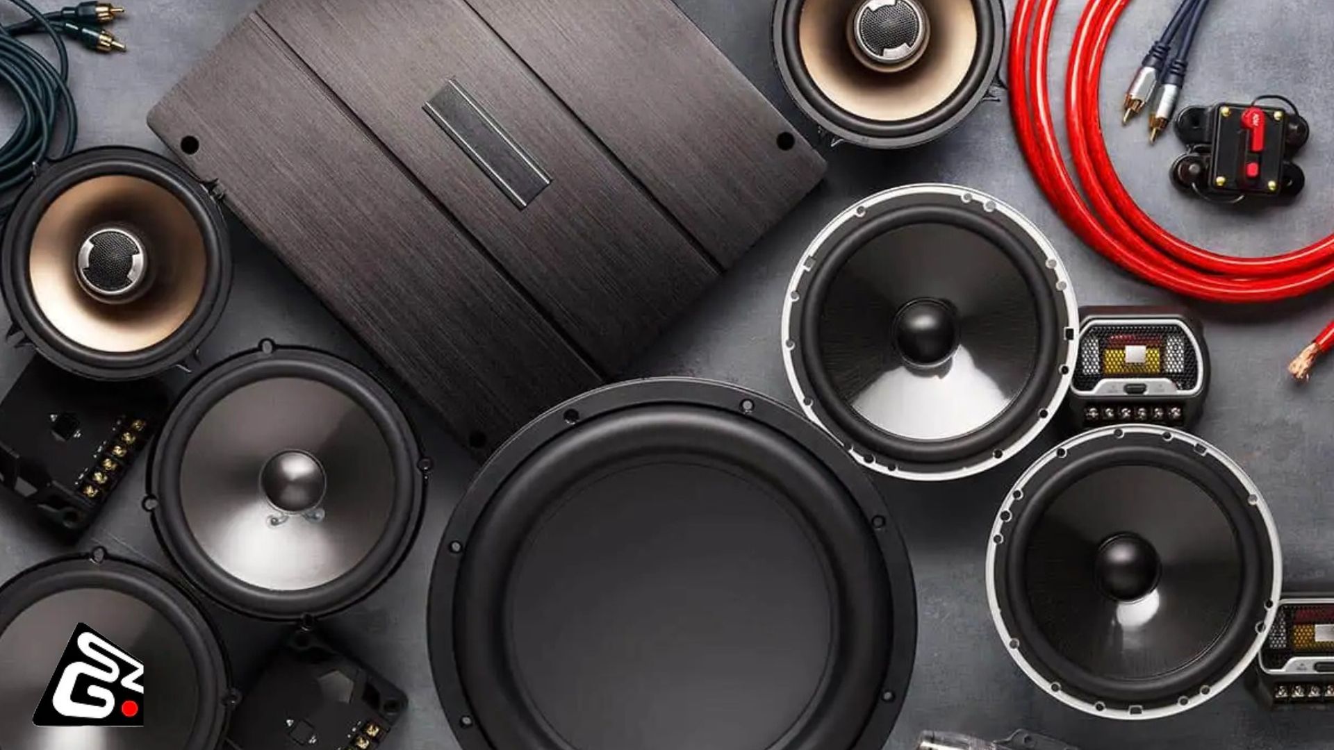 Flat lay of car audio components including subwoofers, midrange speakers, tweeters, an amplifier, RCA cables, and a fuse holder, all arranged on a dark surface. SG logo is visible.