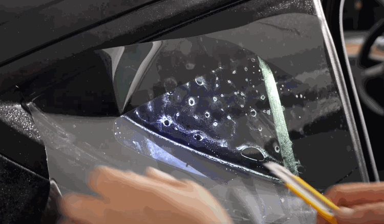 A technician smooths and trims a sheet of tint film on a wet car window using a precision blade.
