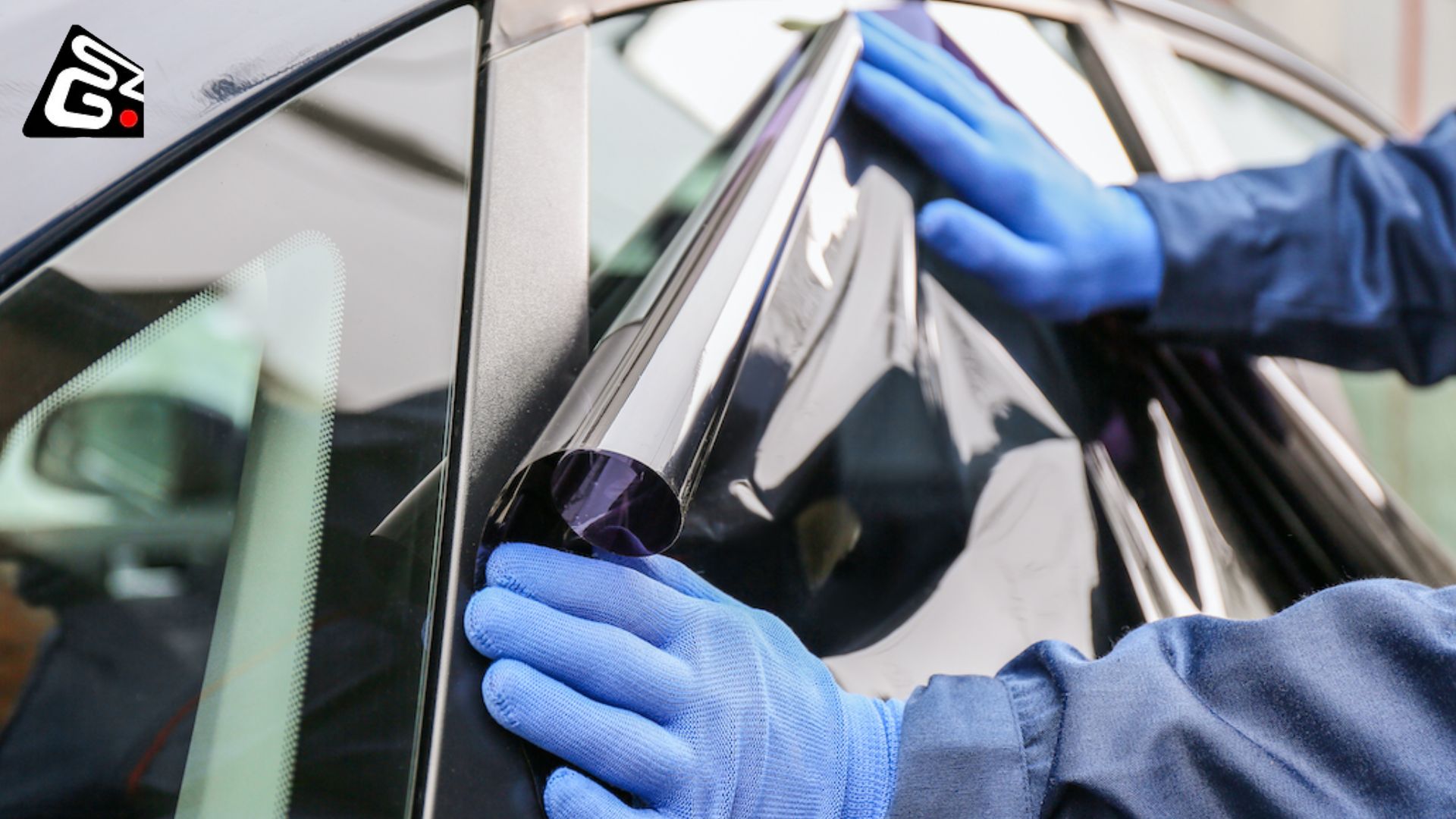 A person wearing blue gloves carefully applies a dark window tint film to the side window of a car.