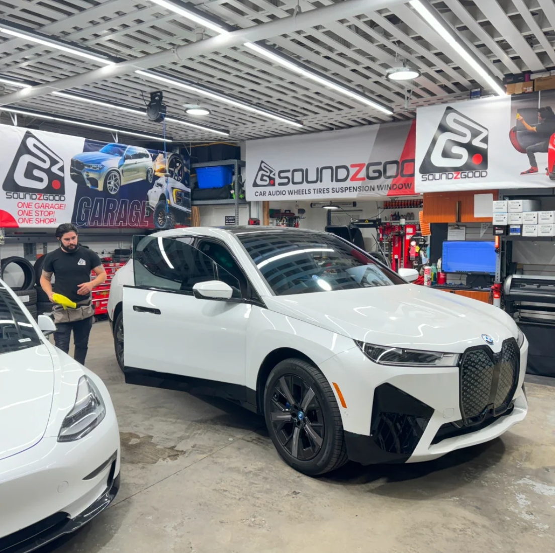 A white BMW iX is parked inside Soundz Good Garage’s workshop in Oxnard, CA, surrounded by custom banners and tools, with a technician nearby preparing for installation or detailing work.
