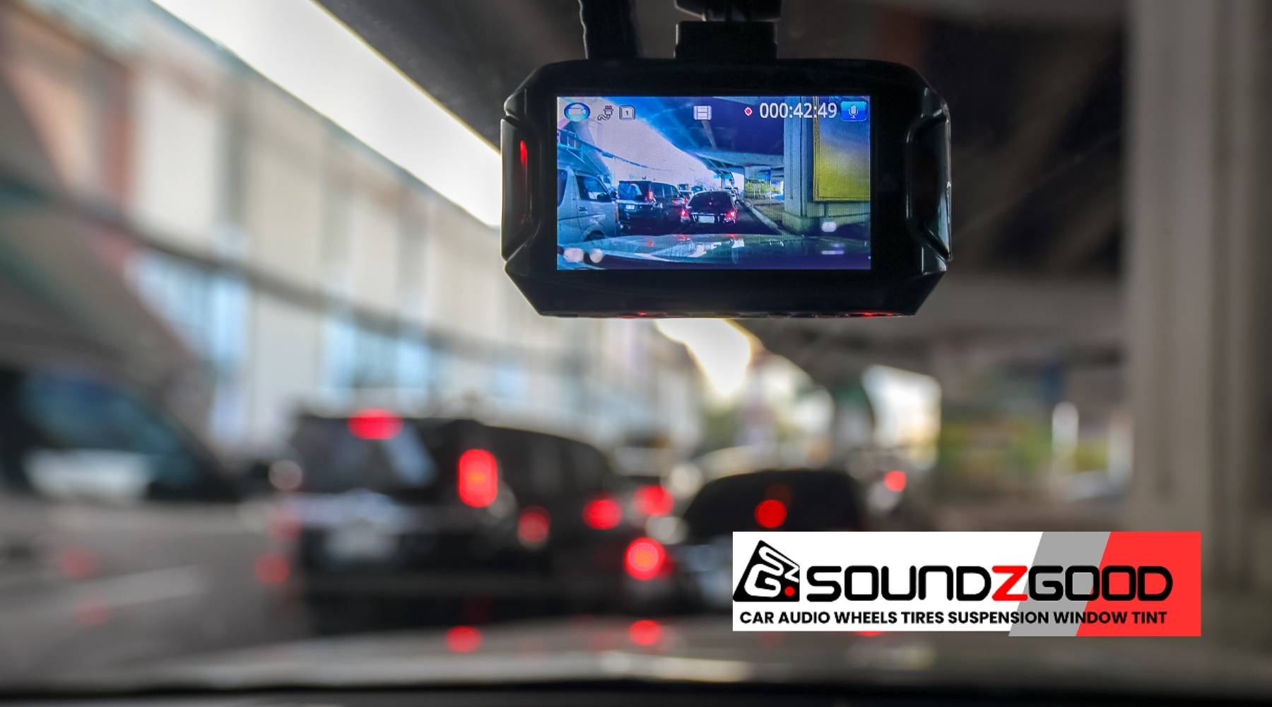 A dash cam recording traffic on a busy city road, showing vehicles ahead through the windshield, with the SoundzGood Garage logo at the bottom.