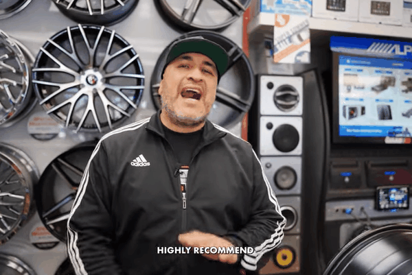 A man wearing a black Adidas jacket and black cap stands inside an automotive shop with car rims displayed on the wall behind him.