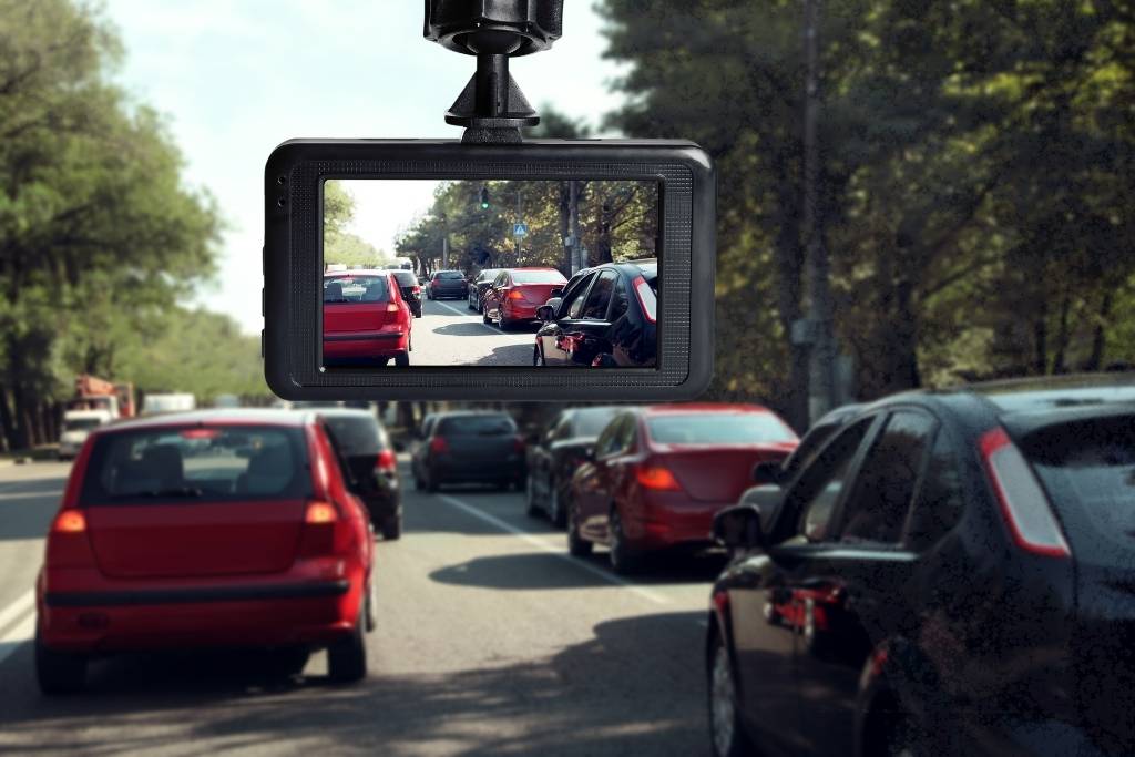 A car dashboard camera records traffic on a busy road lined with trees during daylight.