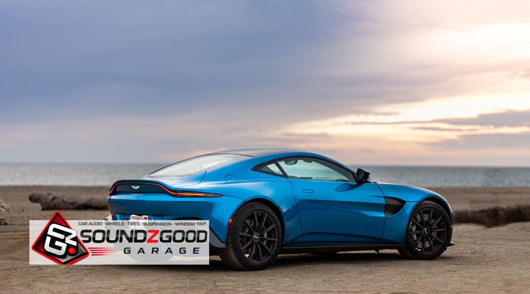 Blue Aston Martin parked by the beach with Soundz Good Garage branding, showcasing a sleek car window tint and custom styling look.