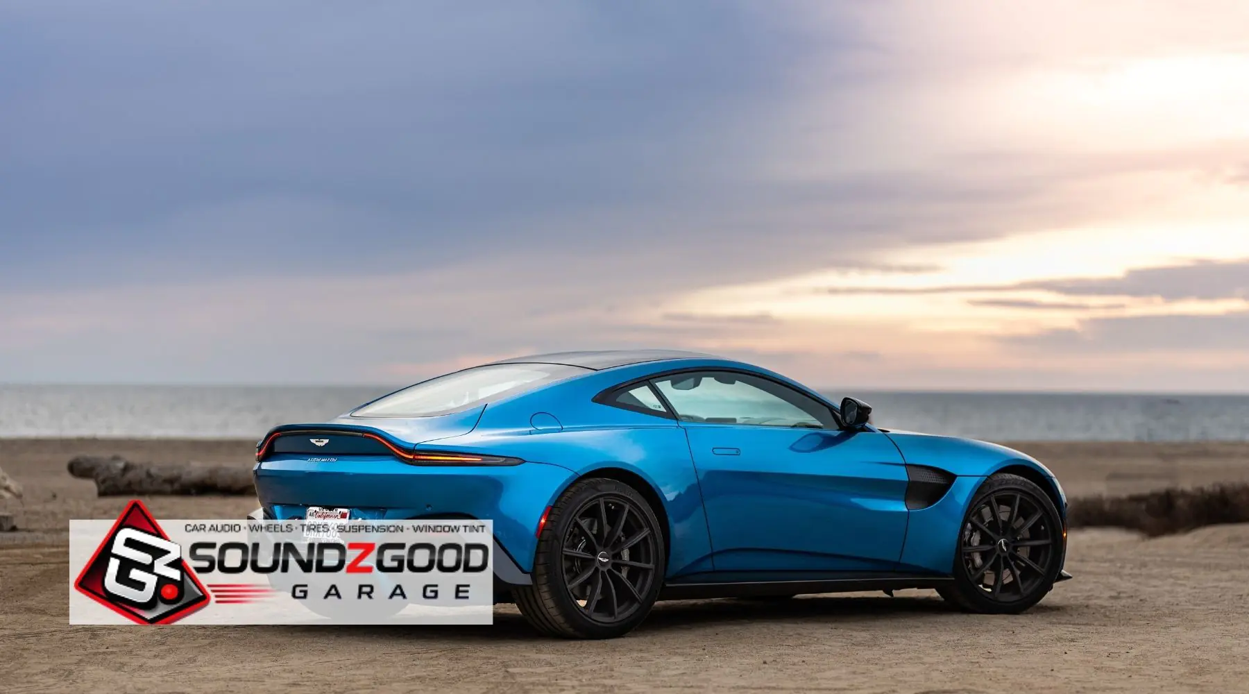 Blue Aston Martin parked by the beach with Soundz Good Garage branding, showcasing a sleek car window tint and custom styling look.