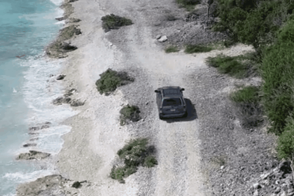 Aerial view of a dark SUV driving along a rocky coastal shoreline beside clear blue water.