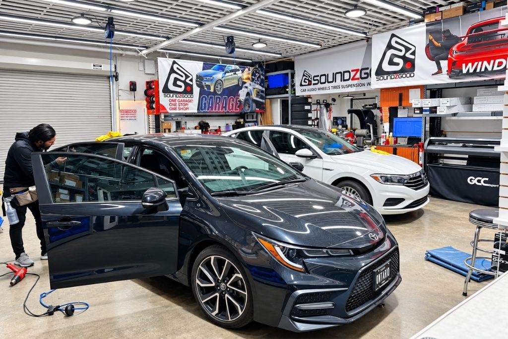 Technician working on a car window inside a Soundz Good Garage automotive shop with multiple vehicles in the service bay.